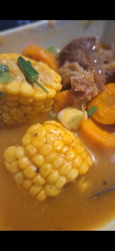 A bowl of stew with corn, carrots, and meat in a flavorful broth.